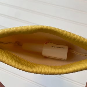 May include: A yellow, textured pouch with a white bottle inside. The bottle has text on it. The pouch is held open, revealing the contents. The interior of the pouch is a lighter yellow.