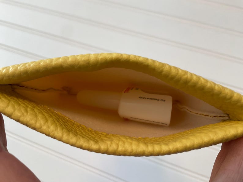 May include: A yellow, textured pouch with a white bottle inside. The bottle has text on it. The pouch is held open, revealing the contents. The interior of the pouch is a lighter yellow.