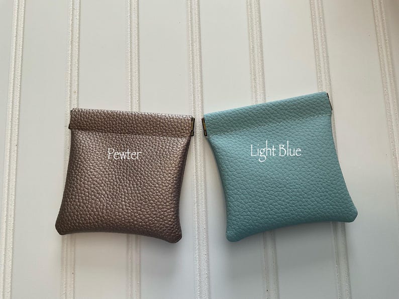 May include: Two small, square pouches in a top-down view. One is pewter-colored with the word "Pewter" embroidered on it. The other is light blue with "Light Blue" embroidered on it. Both pouches have a textured surface.