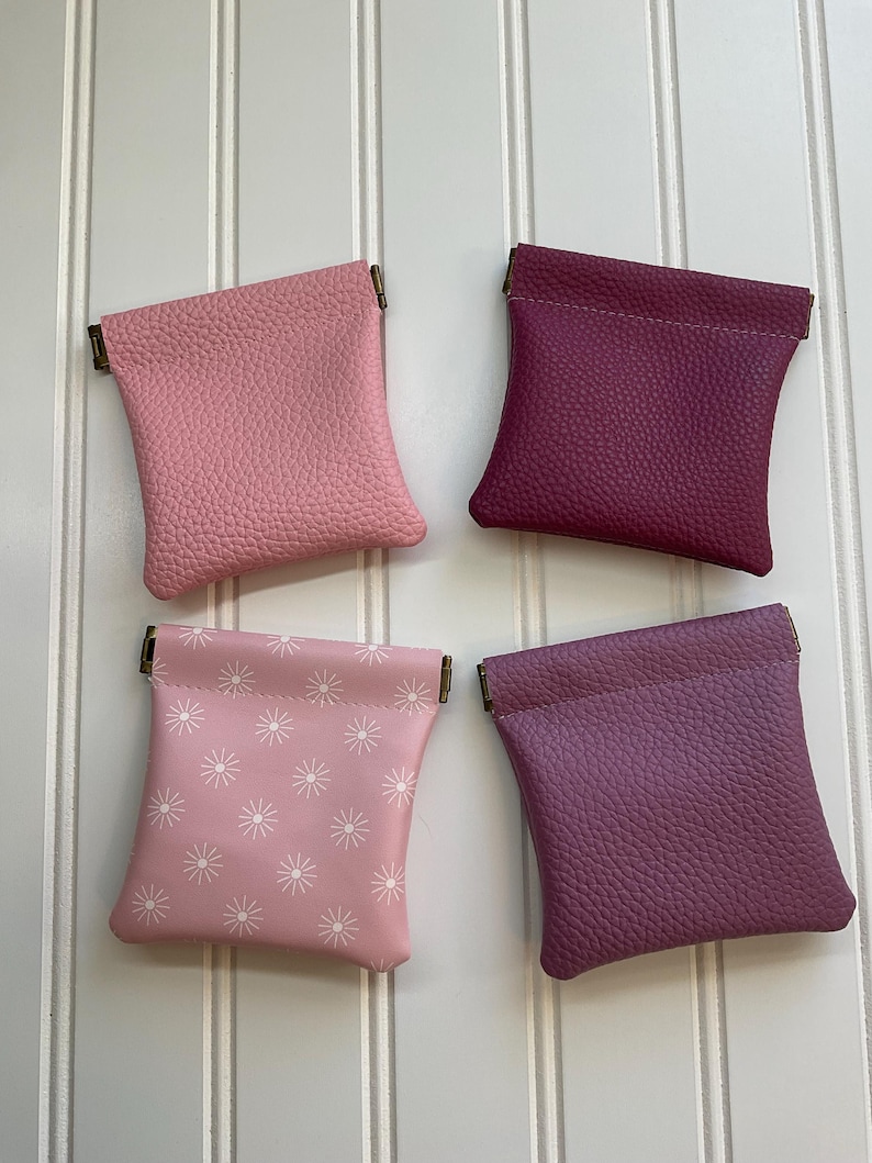 May include: Four small, leather coin purses in shades of pink and purple. Two purses are solid colors, while the other two feature a white starburst pattern. Each purse has a metal clasp.