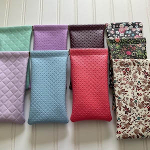 May include: A collection of eyeglass cases in various colors and patterns. The cases include solid colors like lavender, blue, and red, as well as floral and textured designs. The cases are rectangular and appear to be made of a soft material.