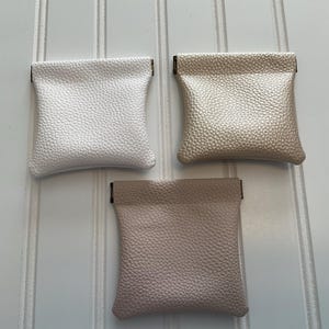 May include: Three small, textured pouches in white, champagne, and blush pink. Each pouch has a rectangular shape with a folded top. The pouches are made of a leather-like material and are displayed against a white background.