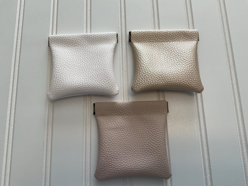 May include: Three small, textured pouches in white, champagne, and blush pink. Each pouch has a rectangular shape with a folded top. The pouches are made of a leather-like material and are displayed against a white background.