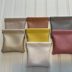 May include: A collection of seven small, textured pouches in various colors, including beige, brown, gray, yellow, pink, and white. Each pouch has a rectangular shape with a folded top and a small metal clasp.