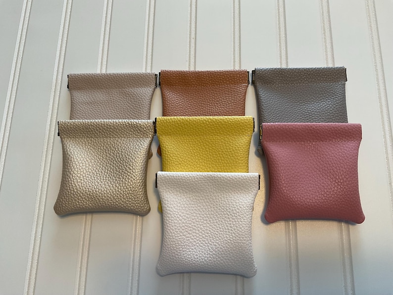 May include: A collection of seven small, textured pouches in various colors, including beige, brown, gray, yellow, pink, and white. Each pouch has a rectangular shape with a folded top and a small metal clasp.