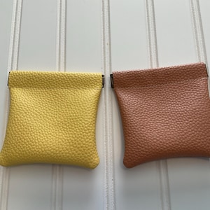 May include: Two small, square coin purses. One is a bright yellow, the other is a muted brown. Both have a textured, pebbled surface and a simple, folded top. The purses are displayed against a white background with vertical lines.