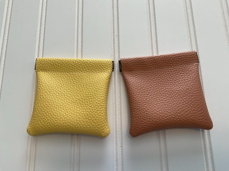 May include: Two small, square coin purses. One is a bright yellow, the other is a muted brown. Both have a textured, pebbled surface and a simple, folded top. The purses are displayed against a white background with vertical lines.