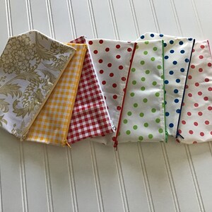 May include: Seven fabric zippered pouches in various patterns. The pouches are lined up in a row, with the top pouch being a white pouch with a gold floral pattern. The second pouch is yellow with a white gingham pattern. The third pouch is red with a white gingham pattern. The fourth pouch is white with red polka dots. The fifth pouch is white with green polka dots. The sixth pouch is white with blue polka dots. The seventh pouch is white with pink polka dots.