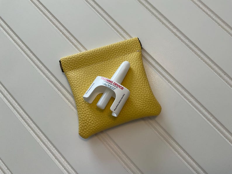 May include: A white training device with the text "TRAINING DEVICE ALTOCO" rests on a yellow textured pouch. The pouch is square-shaped with a black trim.