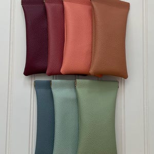 May include: A collection of seven rectangular pouches with a pebbled texture, arranged in two rows. The top row features deep purple, burgundy, coral, and tan colors, while the bottom row displays slate blue, sage green, and light olive green shades.
