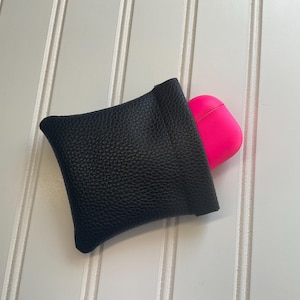 May include: A black leather pouch with a textured surface, holding a bright pink AirPod case. The pouch is square-shaped and has a simple, minimalist design. The background is a white surface with vertical lines.