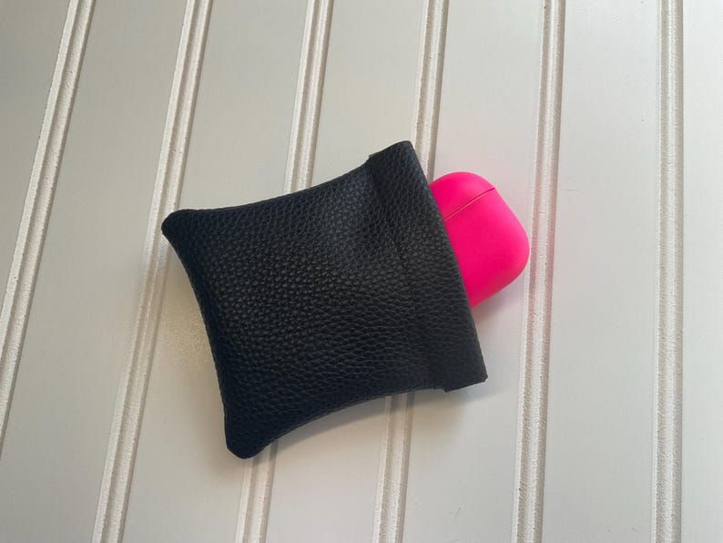May include: A black leather pouch with a textured surface, holding a bright pink AirPod case. The pouch is square-shaped and has a simple, minimalist design. The background is a white surface with vertical lines.