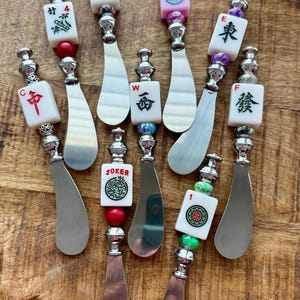 May include: A collection of decorative cheese spreaders with silver blades and ornate handles. Each spreader features a different Mahjong tile design, including "Joker" and various Chinese characters, with colorful bead accents. The spreaders are displayed on a wooden surface.