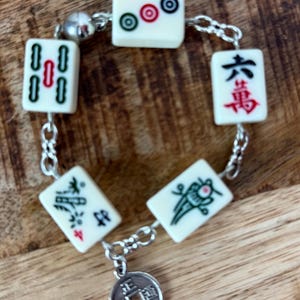 May include: A bracelet featuring ivory-colored mahjong tiles with red, green, and blue markings, connected by a silver chain. A small, round charm with Chinese characters hangs from the bracelet. The bracelet is displayed on a wooden surface.
