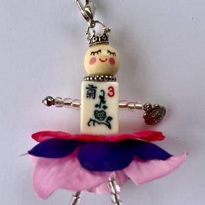 K&ouml;nnte beinhalten: Ein kleiner Puppenanh&auml;nger mit einem rosa-violetten Blumenrock, einem wei&szlig;en Gesicht mit geschlossenen Augen, einer Krone und einer wei&szlig;en Fliese mit chinesischen Schriftzeichen und der Zahl 3.