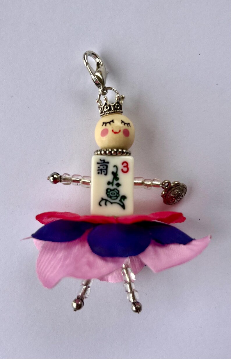 K&ouml;nnte beinhalten: Ein kleiner Puppenanh&auml;nger mit einem rosa-violetten Blumenrock, einem wei&szlig;en Gesicht mit geschlossenen Augen, einer Krone und einer wei&szlig;en Fliese mit chinesischen Schriftzeichen und der Zahl 3.