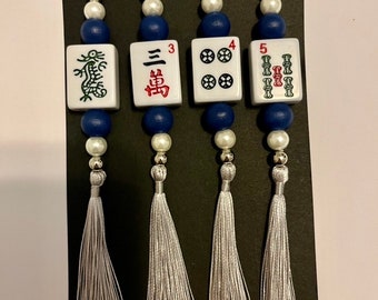 May include: Four white and blue Chinese mahjong tiles with silver tassels hanging from a white string. The tiles feature a dragon, the number 3, the number 4, and the number 5.