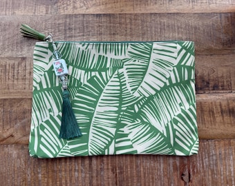 Puede incluir: Bolso de mano rectangular con un estampado de hojas tropicales verdes y blancas. El bolso tiene una cremallera verde y una borla verde unida a un charm de cuentas. El charm tiene la palabra "SPRING".