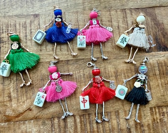 May include: A collection of handmade doll charms with beaded heads, wire arms and legs, and fringed skirts in various colors. Each doll holds a small tile with Chinese characters. The dolls are displayed on a wooden surface.