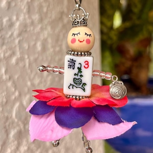 K&ouml;nnte beinhalten: Eine Gl&uuml;cksbringerkette mit einer Puppe mit einem Blumenrock, einer Krone und einem Mahjong-Stein mit der Zahl 3 und dem chinesischen Schriftzeichen "清" (ausgesprochen "qing").