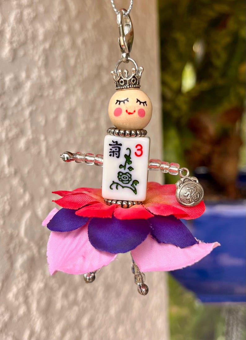 K&ouml;nnte beinhalten: Eine Gl&uuml;cksbringerkette mit einer Puppe mit einem Blumenrock, einer Krone und einem Mahjong-Stein mit der Zahl 3 und dem chinesischen Schriftzeichen "清" (ausgesprochen "qing").