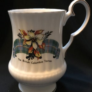 May include: White ceramic mug with a gold rim and handle. The mug features a floral design with a yellow flower and green and blue tartan ribbon. The text "The British Columbia Tartan" is printed below the design.