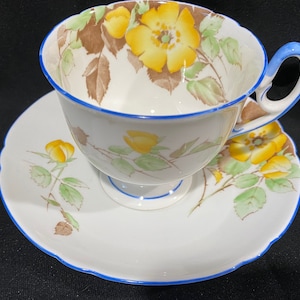Pode incluir: Um conjunto de chávena e pires branco com guarnição azul e padrão floral amarelo. A chávena tem uma pega e o pires tem uma borda ondulada.