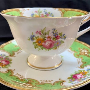 Puede incluir: Una taza de té y un platillo blancos con un patrón floral verde y dorado. La taza tiene un borde festoneado y un asa delicada. El platillo tiene un diseño floral a juego y un borde dorado.