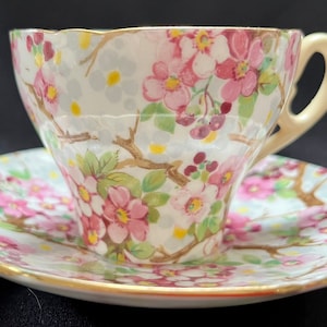 Puede incluir: Juego de taza y platillo floral. La taza y el platillo de porcelana blanca están decorados con flores rosas, hojas verdes y ribetes dorados. La taza tiene un asa color crema y un borde festoneado. El platillo también tiene un borde festoneado.