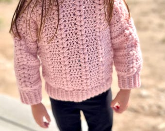 May include: A light pink, hand-crocheted sweater with a textured stitch pattern. The sweater features a crew neck and ribbed cuffs and hem. The sleeves are long, and the garment is made of soft yarn. The sweater is designed for children.