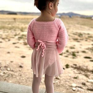 Puede incluir: Una niña pequeña con un top rosa de ganchillo y una falda de ballet. El top de manga larga tiene un escote cuadrado y se ata a la cintura. Lleva una falda de ballet rosa a juego, medias y zapatos brillantes. Un lazo rosa adorna su cabello.