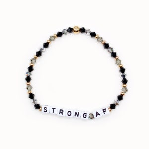 May include: A black and gray crystal beaded bracelet with a gold clasp. The bracelet has white letter beads that spell out "STRONG AF".