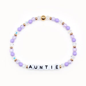 May include: A purple and clear beaded bracelet with a gold accent and the word "AUNTIE" spelled out in white letters.