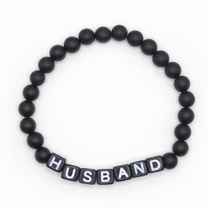 May include: Black beaded bracelet with the word "HUSBAND" spelled out in white letters.