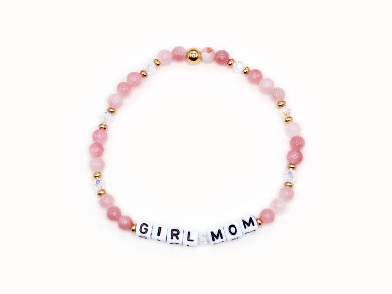 K&ouml;nnte beinhalten: Ein rosa-wei&szlig;es Perlenarmband mit goldenen Akzenten und wei&szlig;en Buchstabenperlen, die "GIRL MOM" buchstabieren.