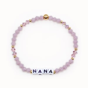 May include: A light purple beaded bracelet with the word "NANA" spelled out in white letter beads. The bracelet features gold accents and is set against a white background. The beads are a mix of faceted and round shapes.