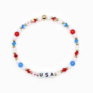 May include: A red, white and blue beaded bracelet with gold accents and the word "USA" spelled out in white letters.
