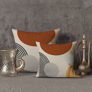 May include: Two decorative throw pillows with abstract geometric designs in rust, black, and beige. A clear glass teacup and a gold-colored metal pitcher are placed next to the pillows, suggesting a home decor theme.