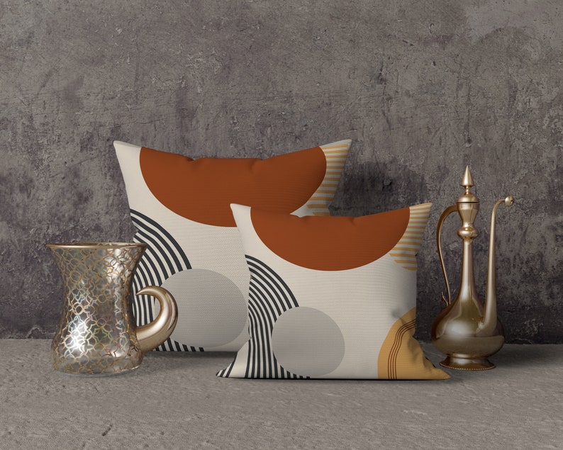 May include: Two decorative throw pillows with abstract geometric designs in rust, black, and beige. A clear glass teacup and a gold-colored metal pitcher are placed next to the pillows, suggesting a home decor theme.