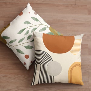 May include: Two decorative throw pillows with abstract designs. One pillow features a branch with green leaves and brown dots on a cream background. The other pillow has a geometric design with brown, yellow, black, and gray shapes on a cream background.