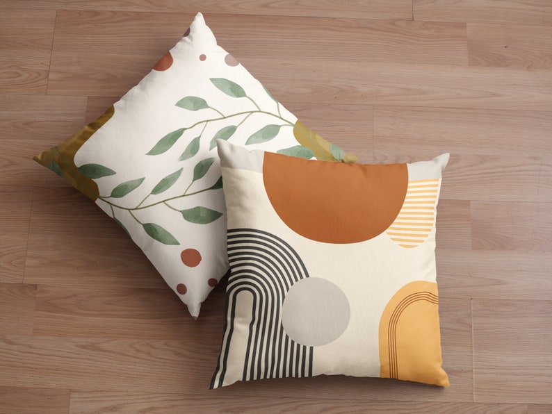 May include: Two decorative throw pillows with abstract designs. One pillow features a branch with green leaves and brown dots on a cream background. The other pillow has a geometric design with brown, yellow, black, and gray shapes on a cream background.