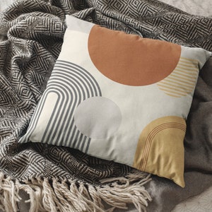 May include: A decorative pillow with a geometric design featuring a series of arches, circles, and stripes in shades of gray, brown, and yellow. The pillow is resting on a gray blanket with a diamond pattern and fringe.