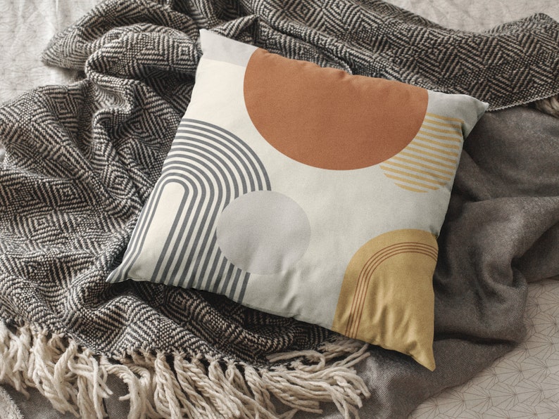 May include: A decorative pillow with a geometric design featuring a series of arches, circles, and stripes in shades of gray, brown, and yellow. The pillow is resting on a gray blanket with a diamond pattern and fringe.