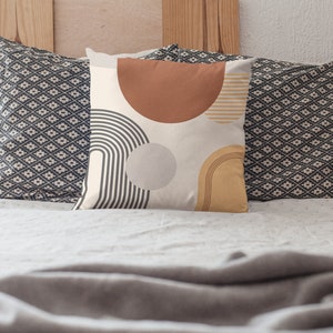 May include: Decorative throw pillow with a cream background and abstract geometric shapes in rust, gray, and mustard yellow. The pillow features a half-circle, a circle, and curved lines. Two patterned pillows are behind it.