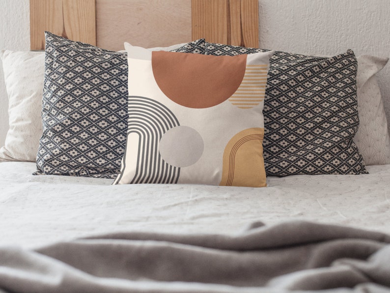 May include: Decorative throw pillow with a cream background and abstract geometric shapes in rust, gray, and mustard yellow. The pillow features a half-circle, a circle, and curved lines. Two patterned pillows are behind it.