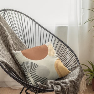 May include: A decorative pillow with a geometric design featuring brown, gray, and yellow shapes on a cream background. The pillow rests on a black wire chair with a patterned throw blanket.