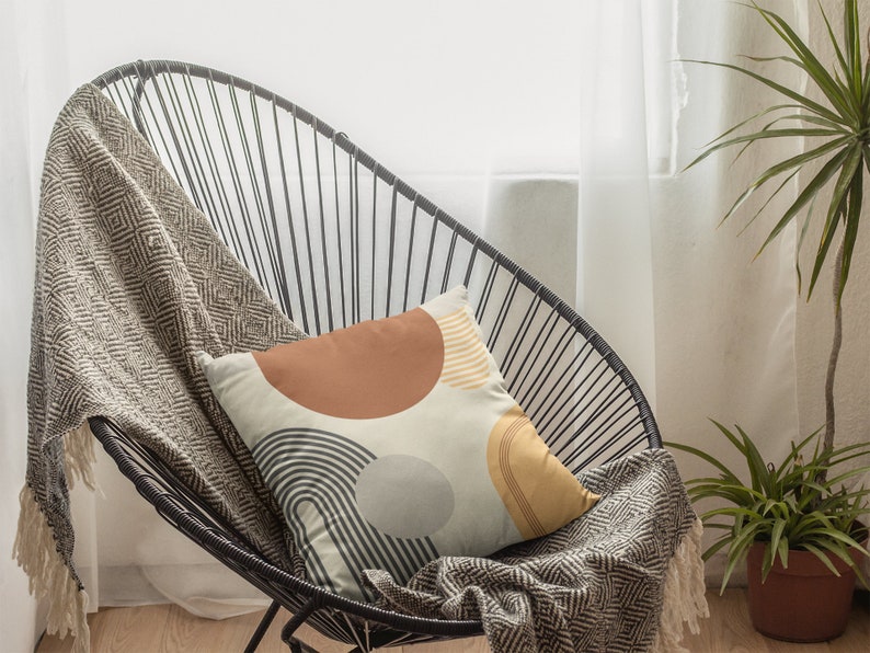 May include: A decorative pillow with a geometric design featuring brown, gray, and yellow shapes on a cream background. The pillow rests on a black wire chair with a patterned throw blanket.