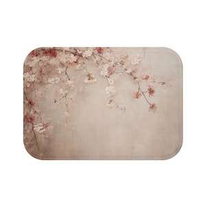 May include: A rectangular bath mat featuring a floral design. The mat has a soft, neutral background with a watercolor-style depiction of cherry blossom branches in shades of pink and white. The mat has rounded corners.