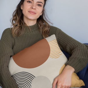 May include: Decorative throw pillow with a cream background and geometric shapes in brown, tan, and gray. The pillow features a large brown semi-circle, tan stripes, and a gray circle and curved lines. The pillow is being held by a person wearing a green sweater.