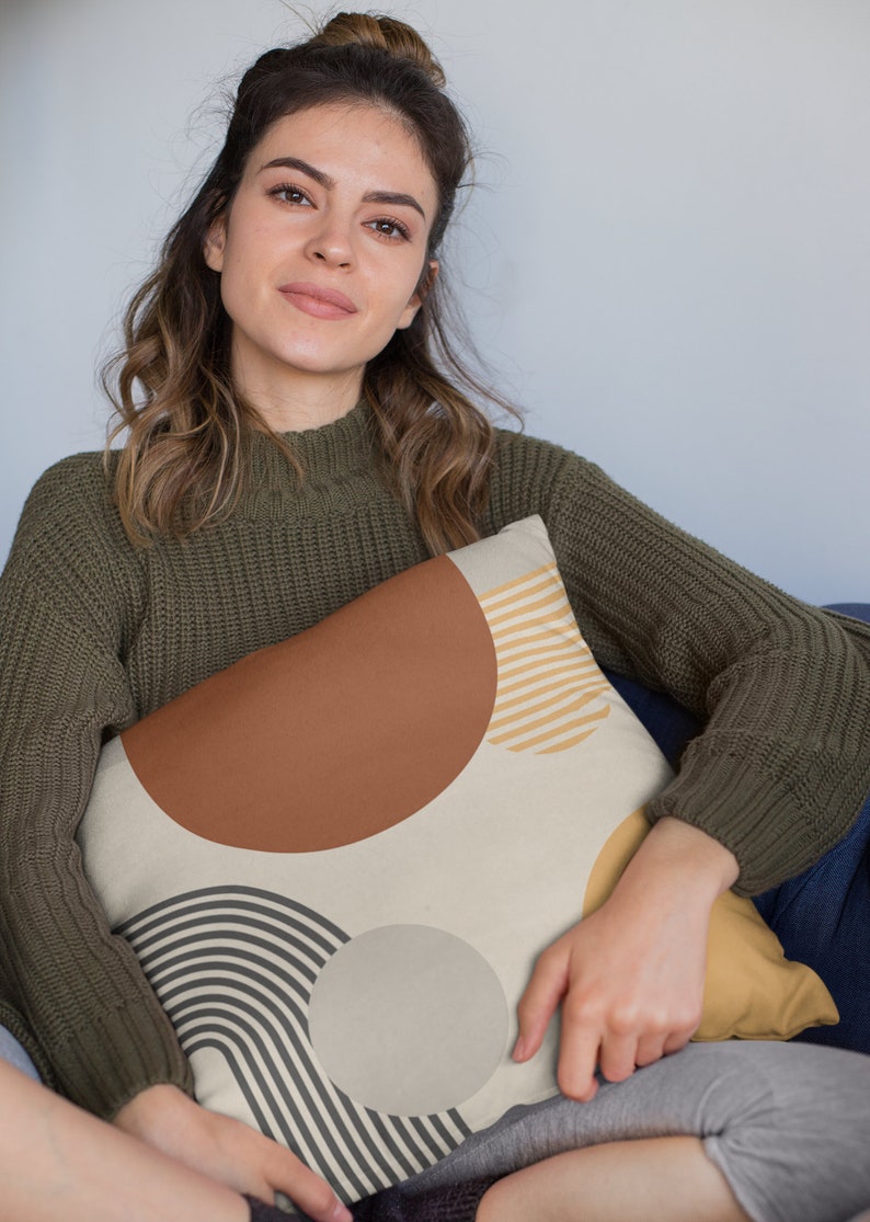 May include: Decorative throw pillow with a cream background and geometric shapes in brown, tan, and gray. The pillow features a large brown semi-circle, tan stripes, and a gray circle and curved lines. The pillow is being held by a person wearing a green sweater.